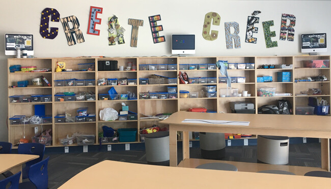 The Create Room, which has the words "Create/Creer" in bigger letters on the wall. Under the letters, there are wooden shelves with different boxes and supplies. In the room, there are tables and, chairs, and round stools.